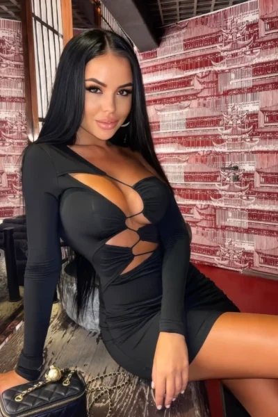 Stylish brunette escort Amsterdam sitting in black dress with long hair and luxury vibe